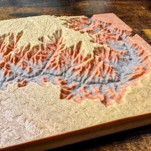 A 3D-printed topographic map in tan, orange, and gray, showing mountain ranges and valleys on a wooden surface.