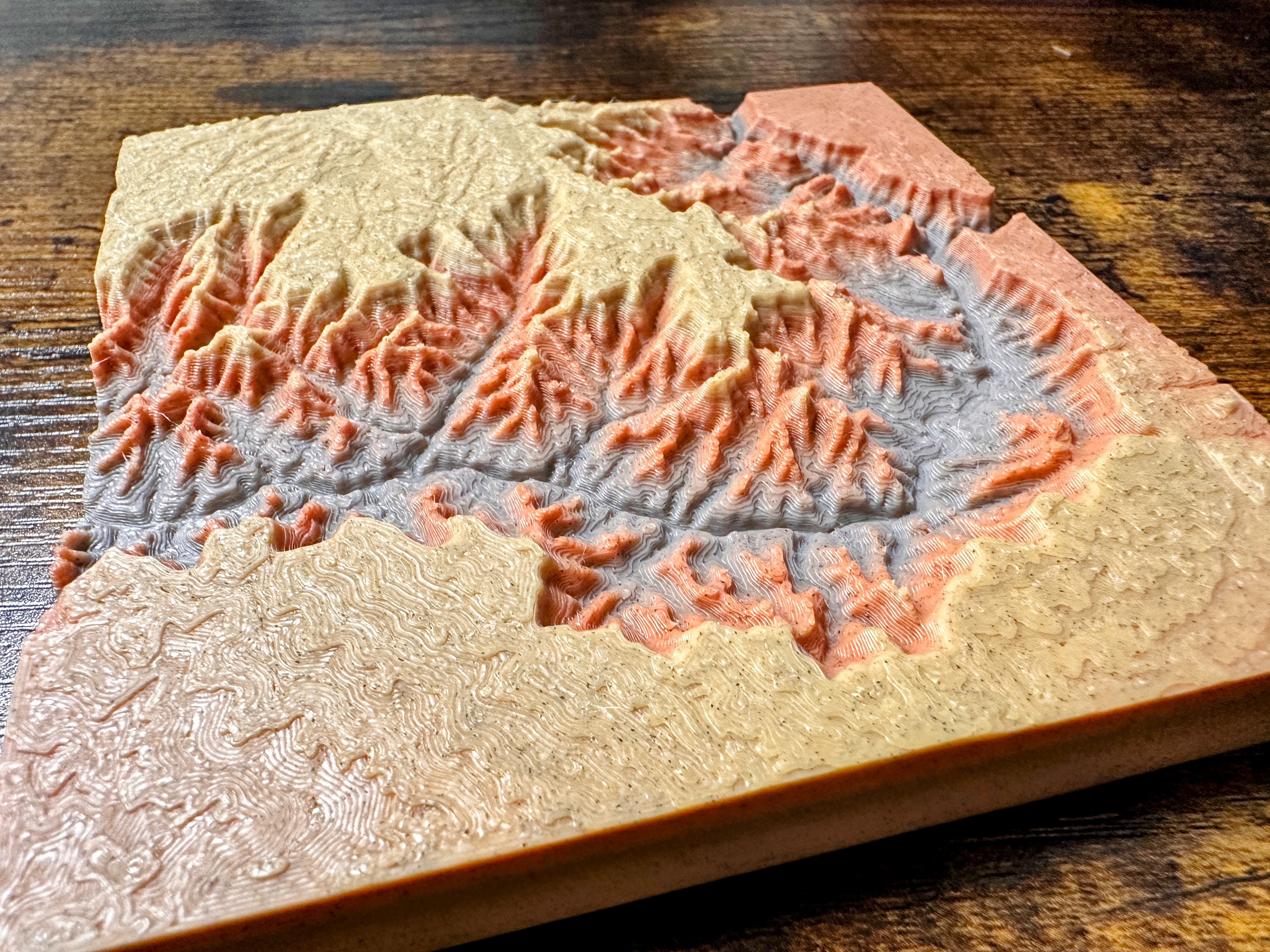 A 3D-printed topographic map in tan, orange, and gray, showing mountain ranges and valleys on a wooden surface.