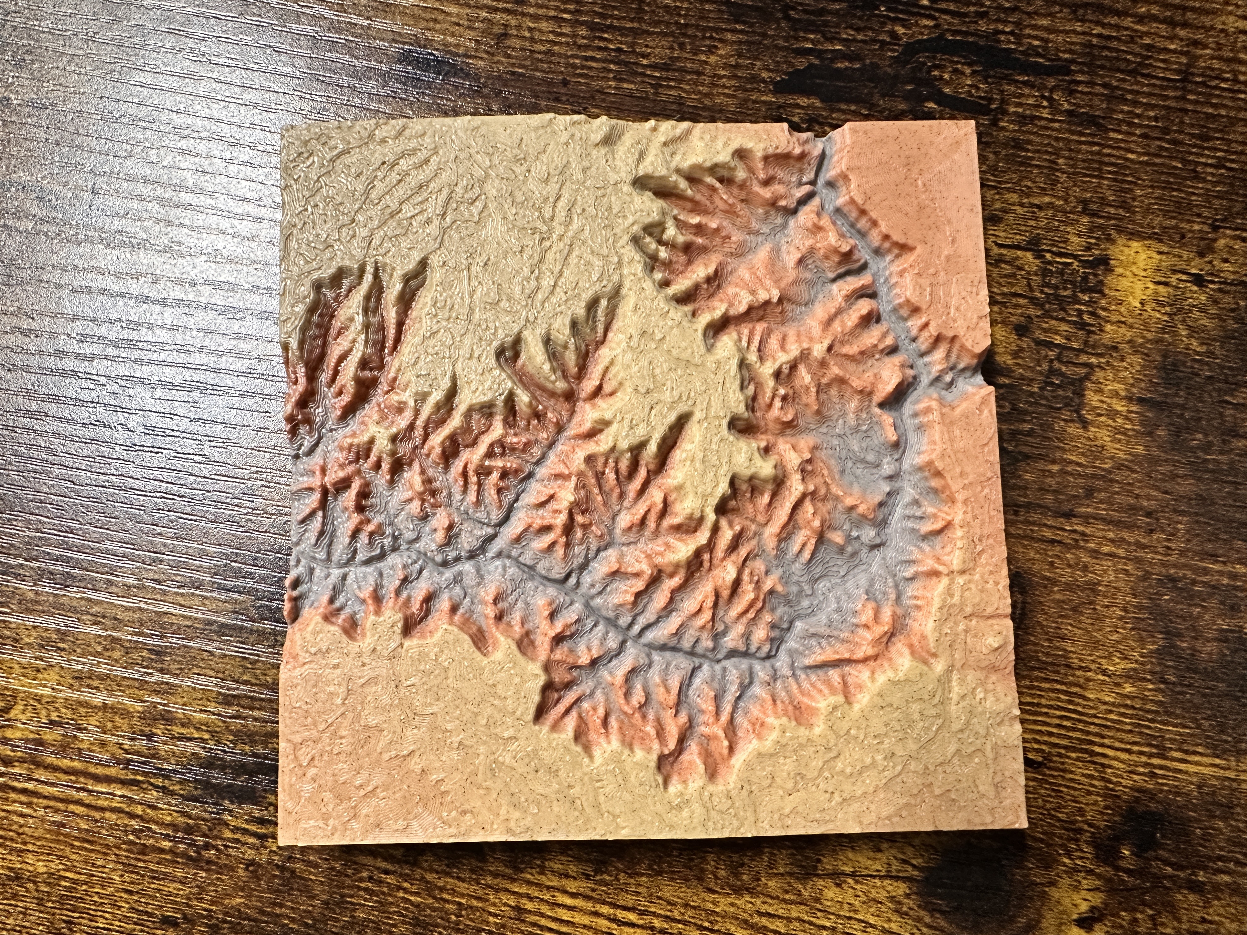 Canyon Ministries Grand Canyon 3D Model A 3D-printed topographic map in tan, orange, and gray displays canyons, ridges, elevation changes, and river paths on wood.