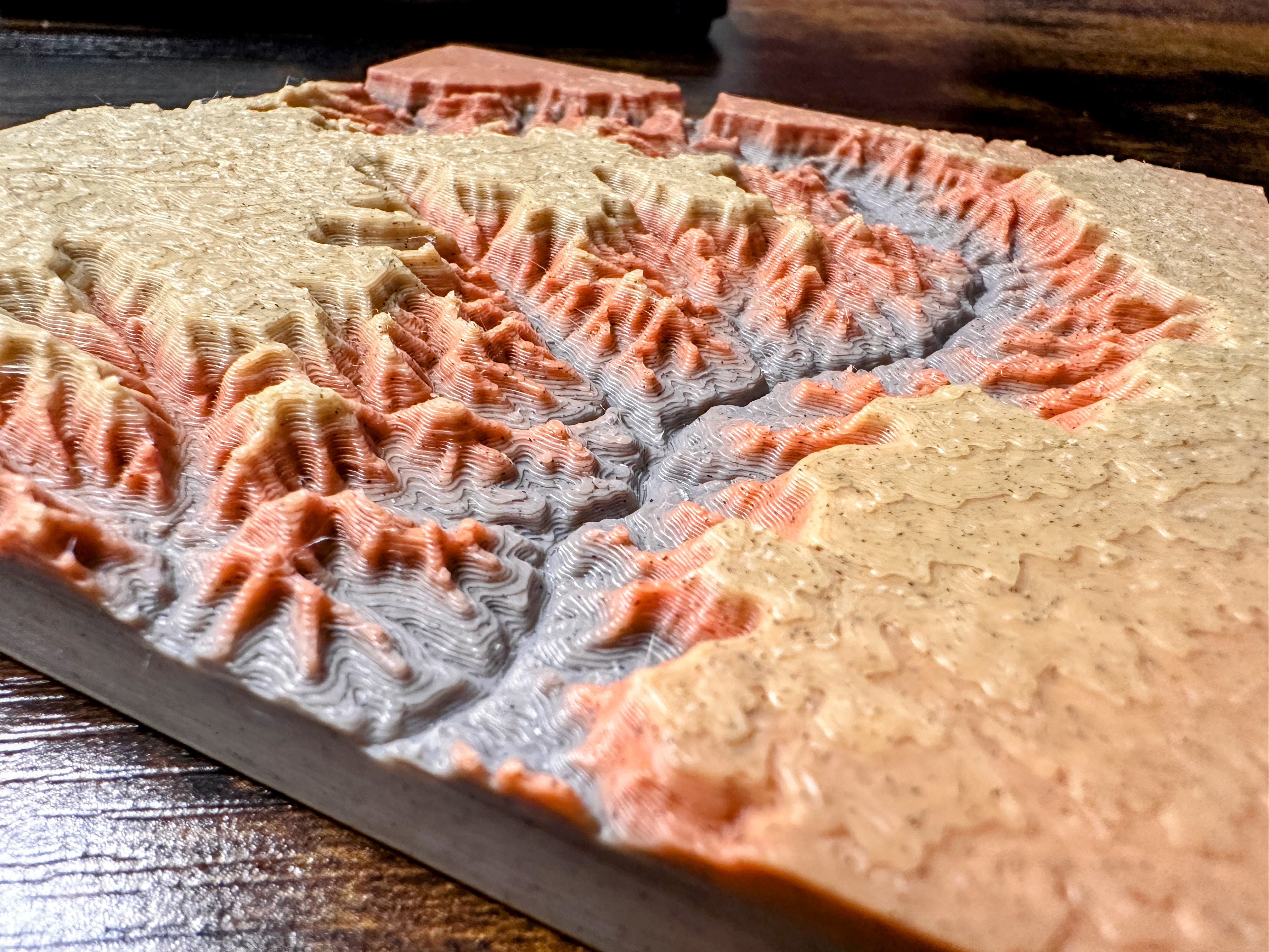 Canyon Ministries Grand Canyon 3D Model A close-up of a 3D-printed topographic map with detailed ridges and valleys in beige, orange, and gray on a wooden surface.