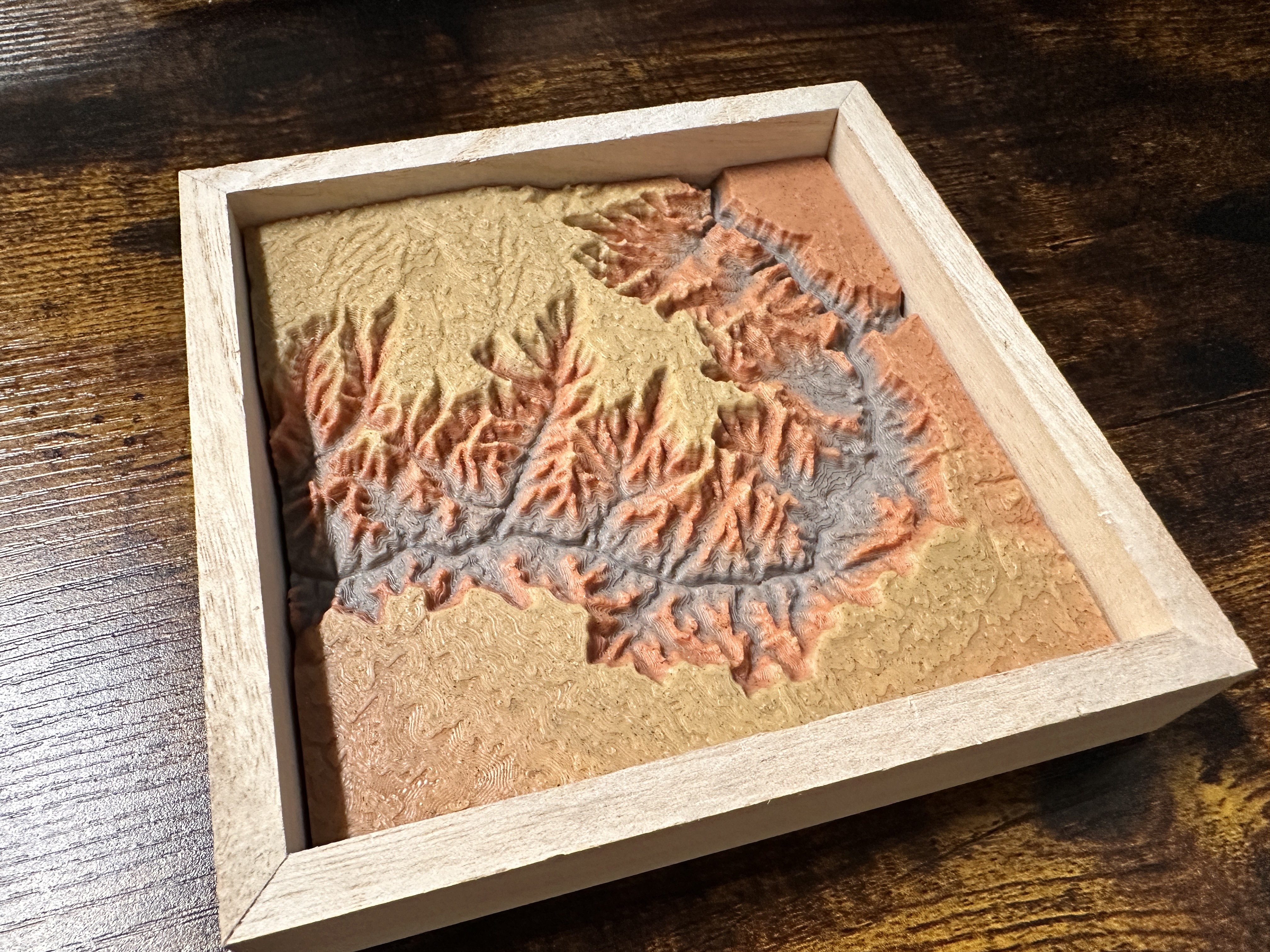Canyon Ministries Grand Canyon 3D Model A wooden frame displays a vivid 3D topographic relief map of mountains, valleys, and rivers in earth tones on a dark wooden table.