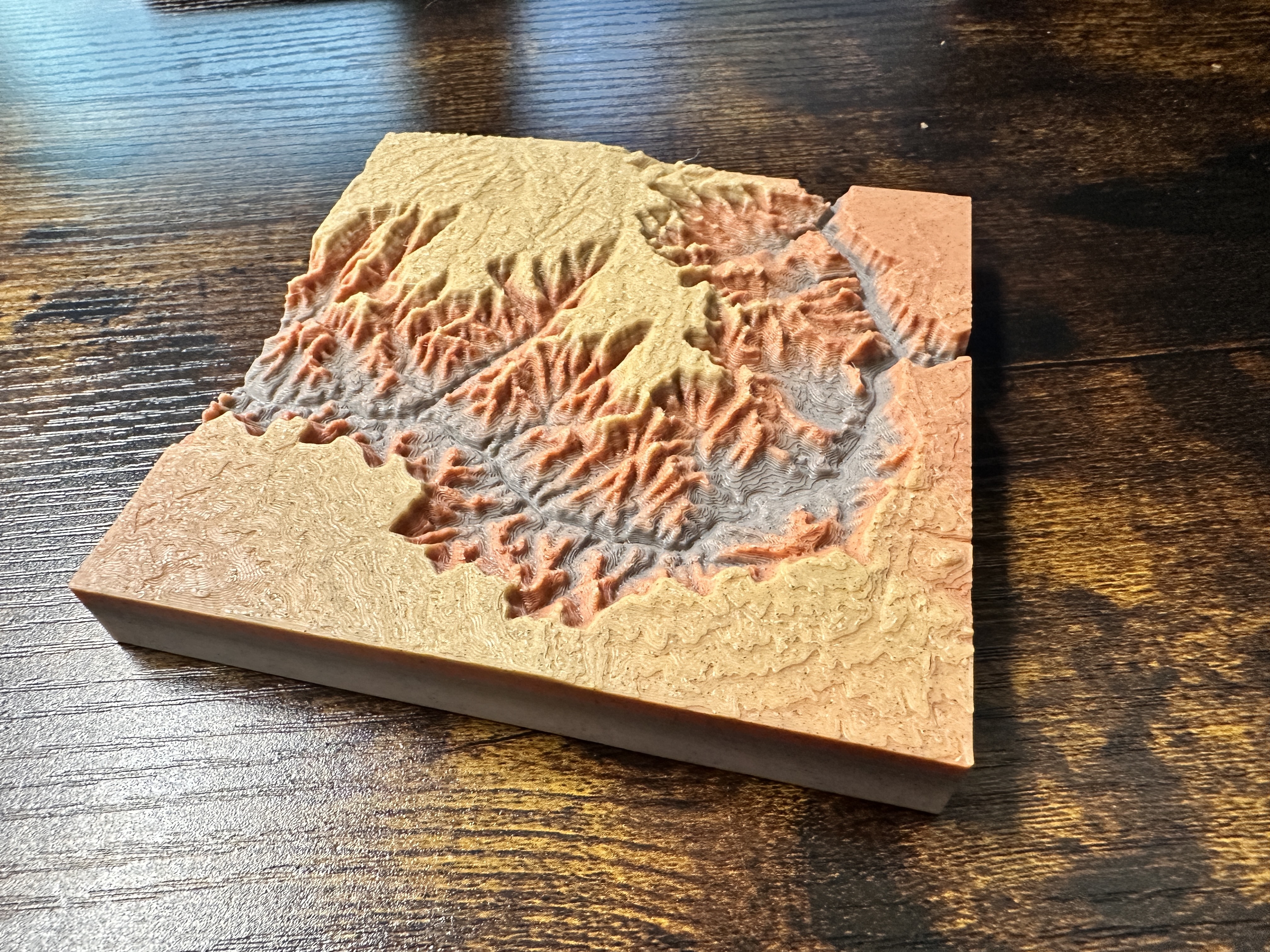 Canyon Ministries Grand Canyon 3D Model A 3D topographical model in earthy tones, shaped like a canyon or mountain range, rests on a dark wooden surface.