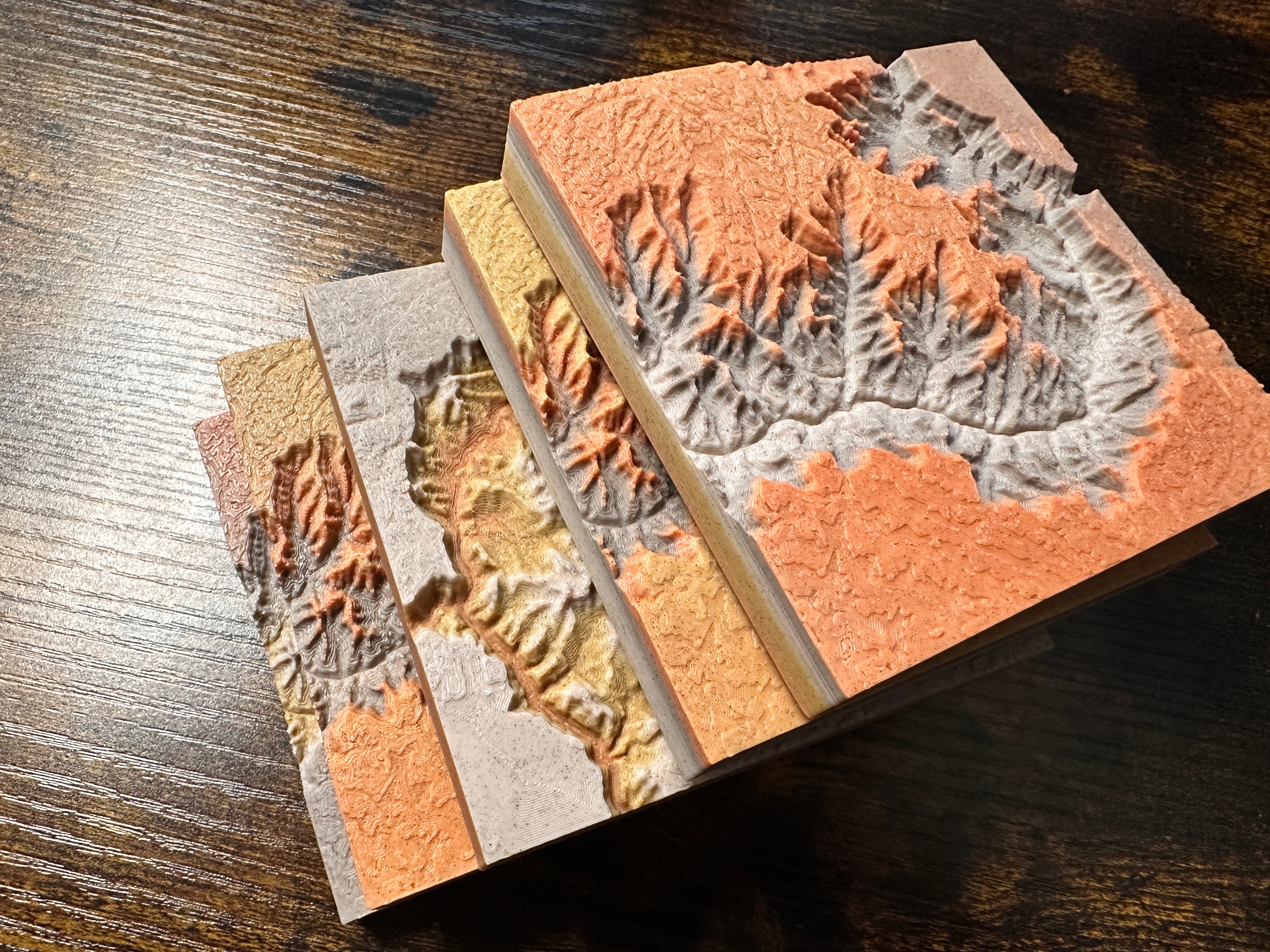 Canyon Ministries Grand Canyon 3D Model Four stacked 3D-printed topographic maps in earthy hues display canyons and ridges on a dark wooden surface.