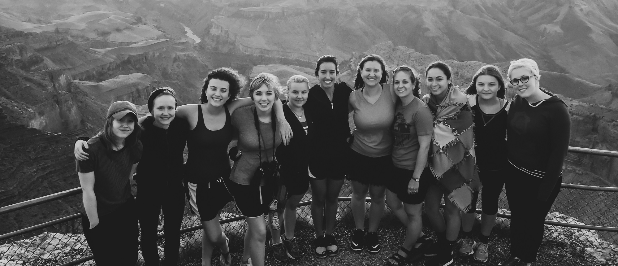 A group of twelve individuals, primarily women, gather closely together, sharing smiles against a striking canyon backdrop during a Grand Canyon group tour. Dressed in casual attire, including athletic wear, they exhibit joyful camaraderie amidst the awe-inspiring scenery.