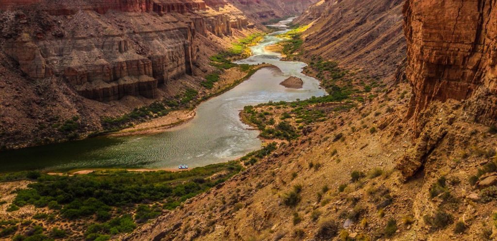 Canyon Ministries offers Grand Canyon River Trips, allowing adventurers to experience the stunning beauty of a winding river cutting through a deep canyon. The steep, layered walls exhibit shades of brown and red, while the riverbanks are dotted with greenery. The glistening water under the sunlight highlights the breathtaking landscape that awaits exploration.
