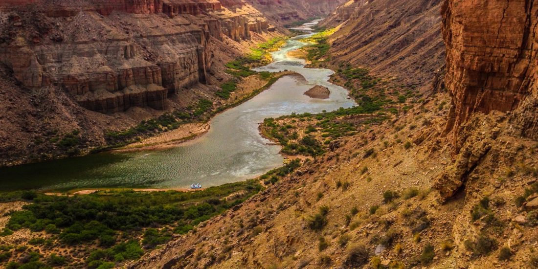 Canyon Ministries offers Grand Canyon River Trips, allowing adventurers to experience the stunning beauty of a winding river cutting through a deep canyon. The steep, layered walls exhibit shades of brown and red, while the riverbanks are dotted with greenery. The glistening water under the sunlight highlights the breathtaking landscape that awaits exploration.