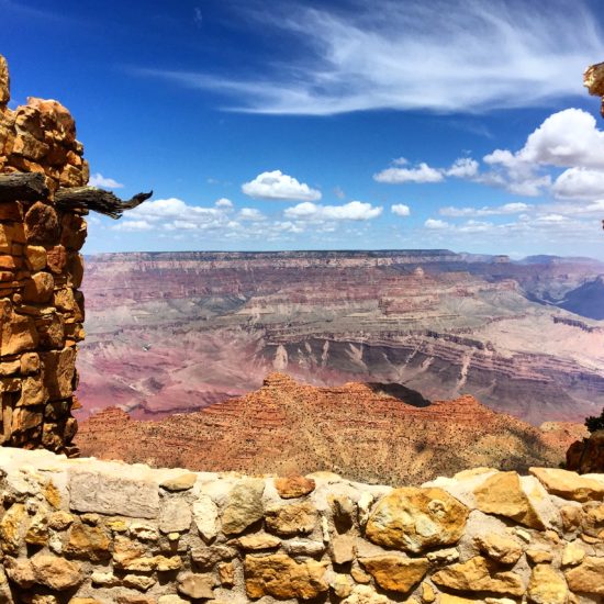 View of the Grand Canyon framed by stone ruins under a partly cloudy blue sky. The rugged canyon landscape, with its red and orange hues, stretches into the distance, creating a dramatic and picturesque scene reminiscent of ancient structures.