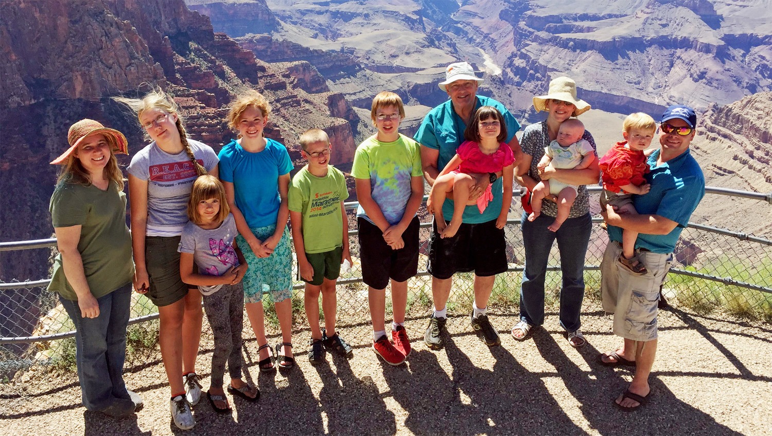 A group of eleven people, a mix of adults and kids, pose cheerfully at a viewpoint overlooking the Grand Canyon on a clear, sunny day—taking part in the Kids Are Free at the Grand Canyon adventure.