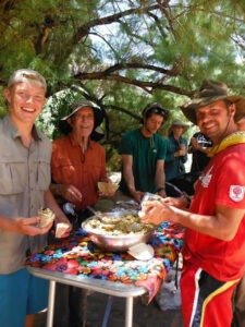 David Losey and several young men standing around a table at mealtime, smiling as they enjoy their food in the shade of the nearby trees