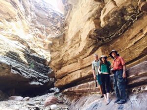 David Losey and two young people stand close together on uneven ground within the canyon, sunlight illuminating the stone walls around them. Two of them shield themselves with hats as they linger amid the arid terrain, a landscape reminiscent of the ancient flood that laid down its stone layers.