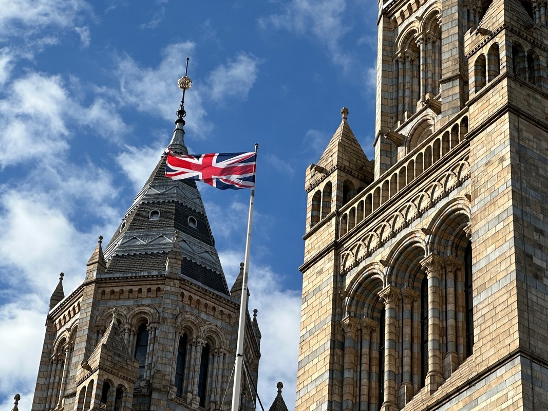A Union Jack flag is prominently displayed on a flagpole before the intricate towers of the Natural History Museum in London. The sky above is blue with scattered clouds, creating a striking backdrop for both the flag and the museums impressive architecture.
