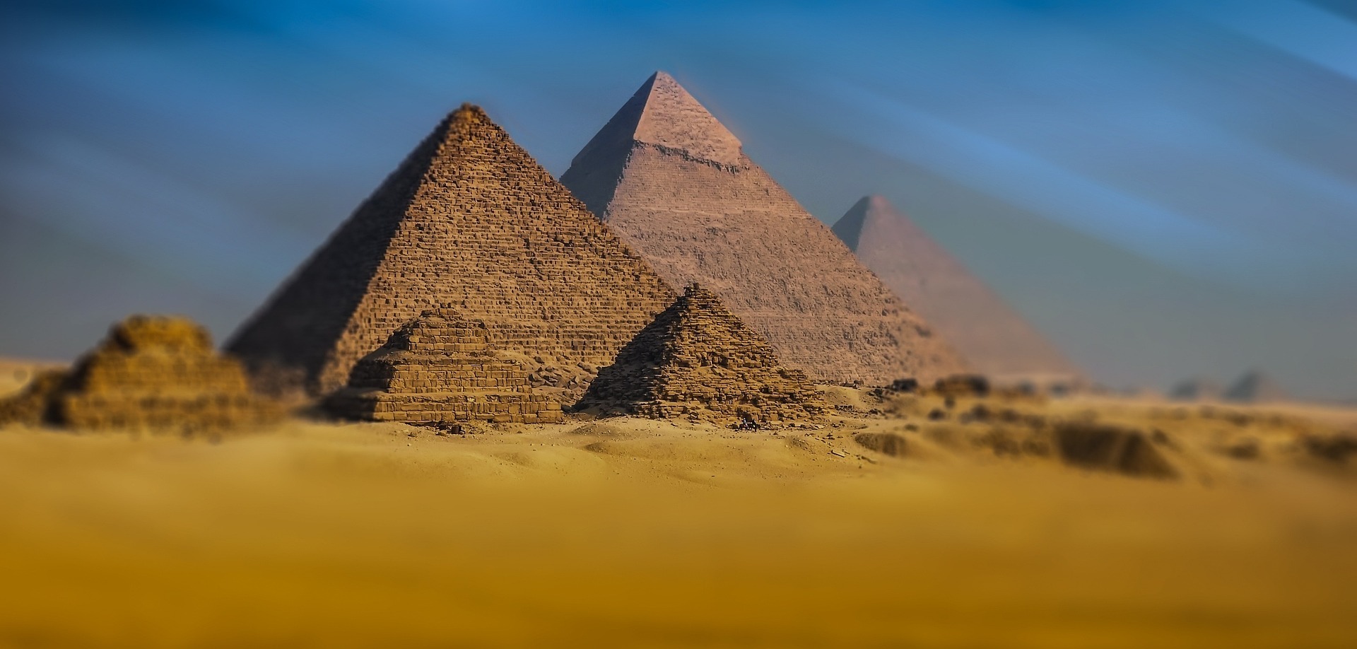 Three large pyramids and several smaller ones rise from a sandy landscape beneath a wide blue sky. The arrangement closely resembles the Pyramids of Giza in Egypt, a renowned location often featured in Egypt Christian Tours. The image is softly focused, particularly around the periphery.