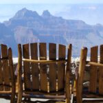 Three people sit side by side on wooden chairs, gazing quietly at the vast expanse of the Grand Canyon’s North Rim. The layered rock formations and misty blue hues create a breathtaking backdrop as they share the tranquil moment together, surrounded by natural beauty under a clear sky.