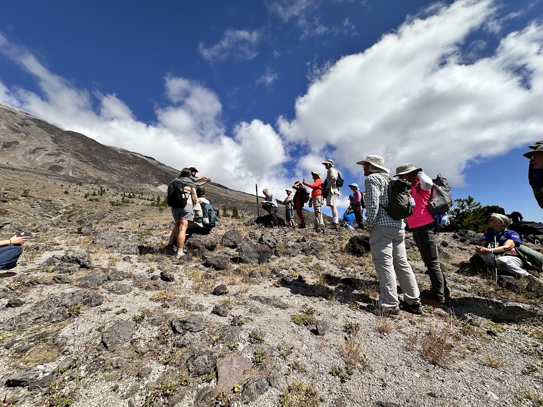 A group of men and women in hats and outdoor clothing walk on rocky, sloped ground under a blue sky near Mount St. Helens.