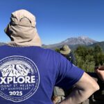 A person in a sun hat and blue Explore Mount St. Helens 45th Anniversary 2025 shirt looks at the mountain as others relax nearby.