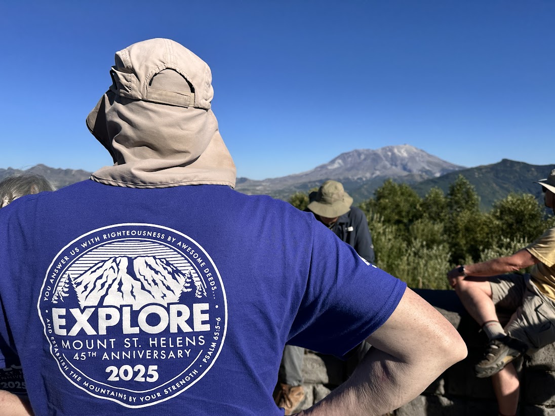 A person in a sun hat and blue Explore Mount St. Helens 45th Anniversary 2025 shirt looks at the mountain as others relax nearby.