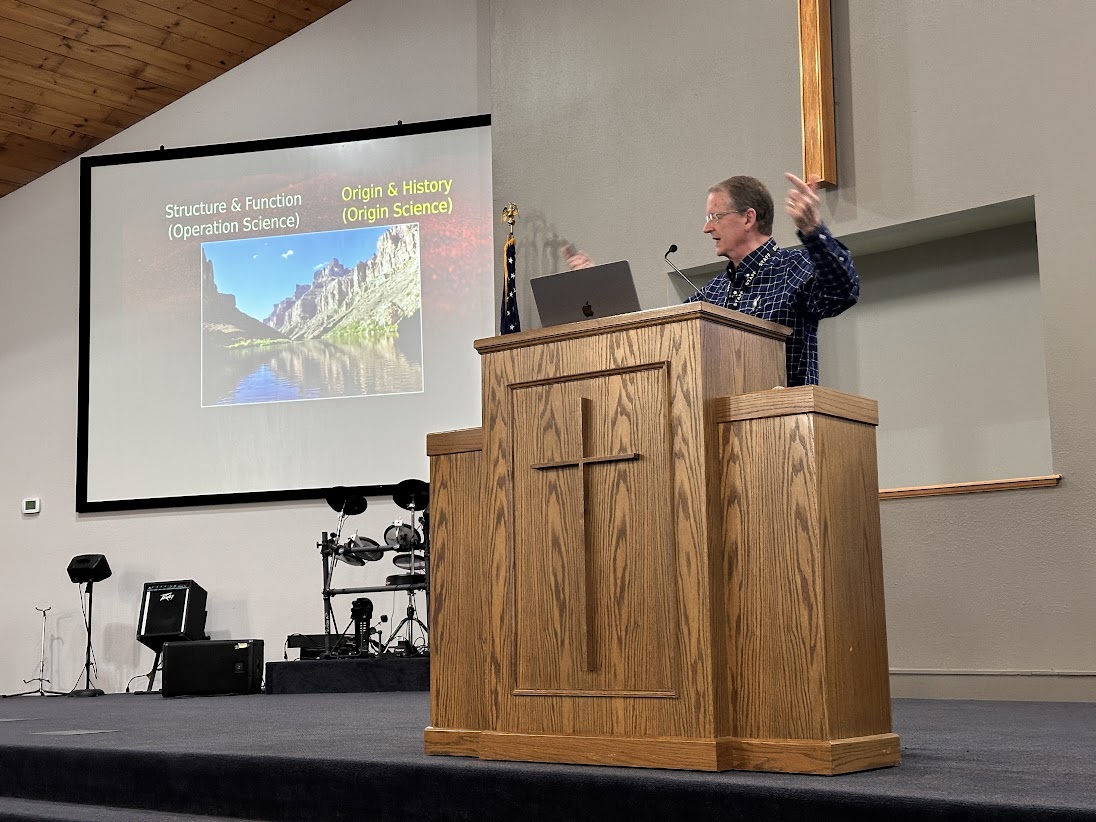 A man stands at a wooden podium with a cross, presenting in a church. Behind him, a slide on Mount St. Helens is displayed.