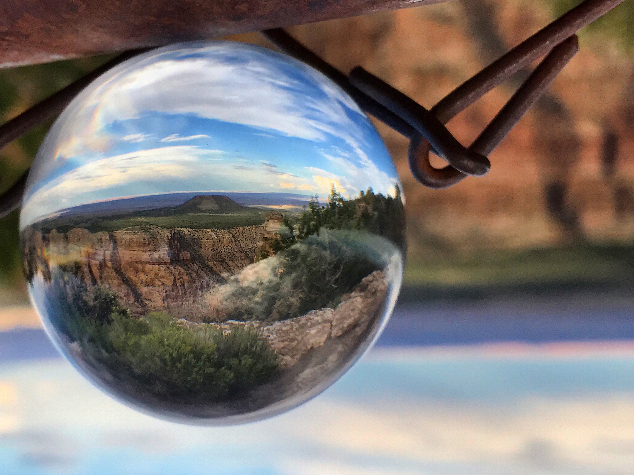 A glass sphere hanging on a wire reflects a flat-topped mountain, blue sky, and greenery in sharp focus; background blurred.