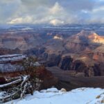 Snow blankets the Grand Canyon’s rocky rim under clouds, sunlight brightening dramatic cliffs and valleys amid wintry pine trees.