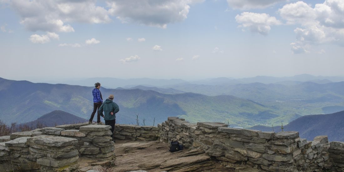 Two people stand on a stone overlook atop a massive mountain, looking at green hills and valleys under a partly cloudy sky. A backpack sits nearby.