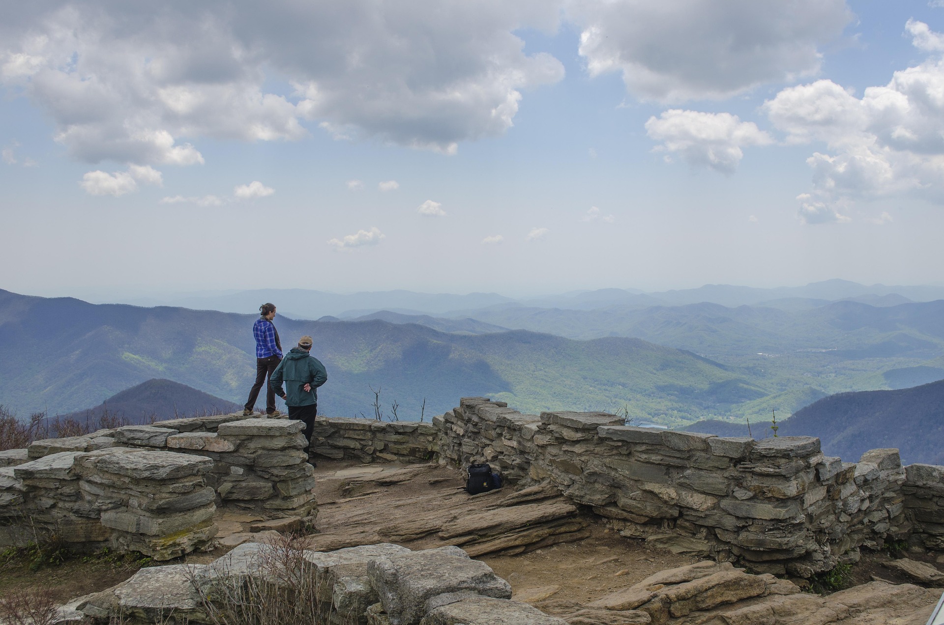 Two people stand on a stone overlook atop a massive mountain, looking at green hills and valleys under a partly cloudy sky. A backpack sits nearby.